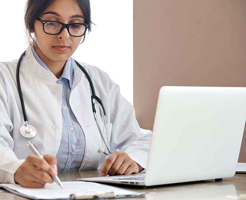 Female doctor sitting in front of laptop and writing with pen on a clipboard.